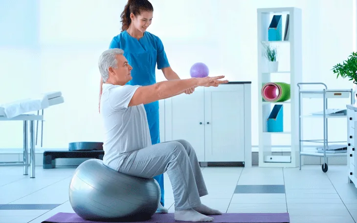 Idoso sentado em uma bola de exercício estendendo os braços, com apoio de uma fisioterapeuta
